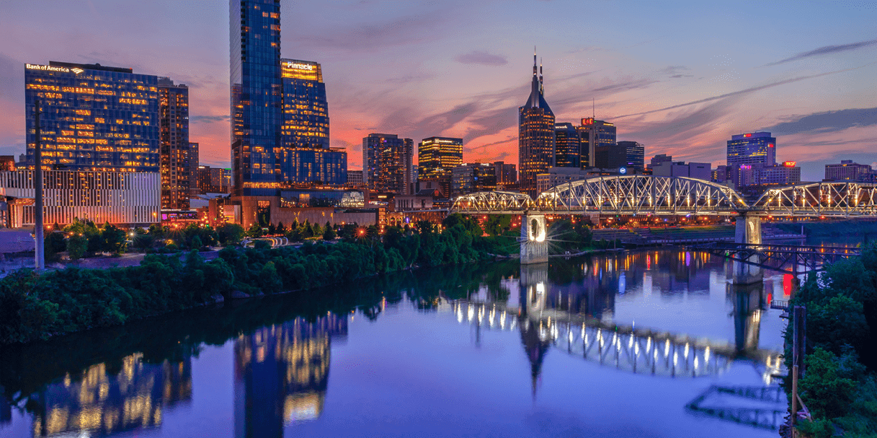 A vibrant view of downtown Nashville, Tennessee, at sunset with illuminated skyscrapers, the iconic AT&T Building, and the John Seigenthaler Pedestrian Bridge reflecting on the calm Cumberland River.